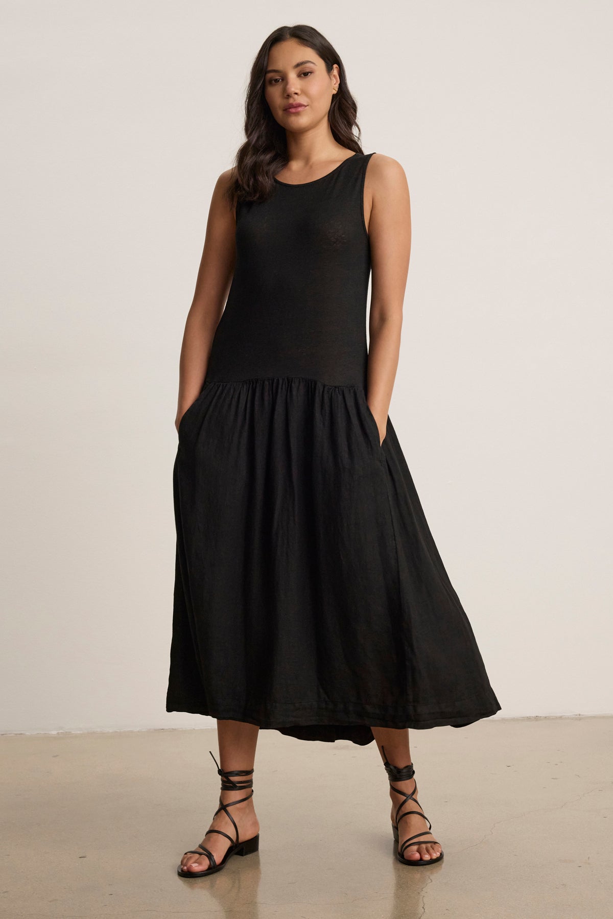 A woman stands against a plain background wearing the Velvet by Graham & Spencer MABLE LINEN KNIT DRESS, a black linen blend tank dress with a flared midi-length skirt and black strappy sandals, her hands in her pockets.