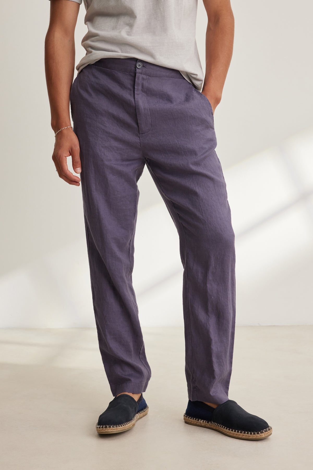 Someone stands indoors with one hand in their pocket, wearing a light grey t-shirt, black espadrilles, and the ALFRED LINEN PANT by Velvet by Graham & Spencer—loose-fitting purple pants featuring an elastic waistband.-39152330571969