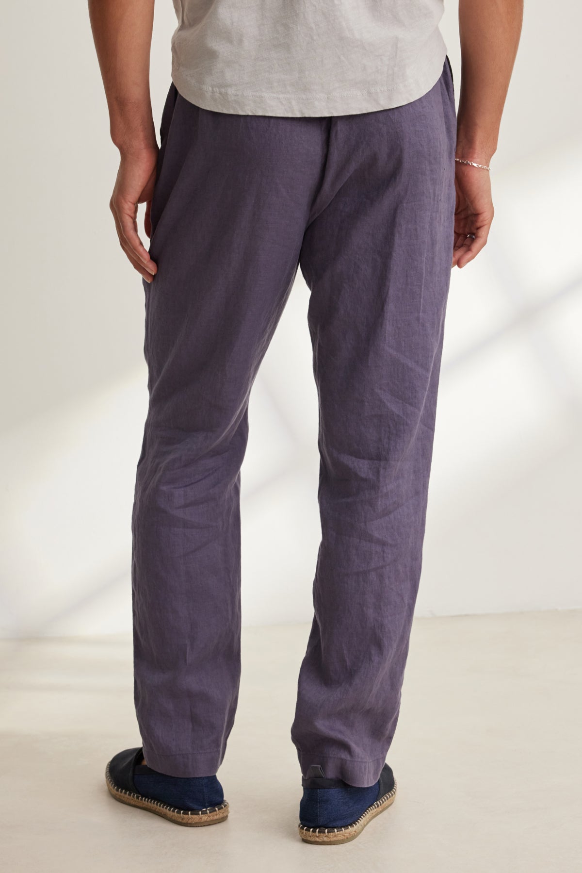 A person stands on a light-colored floor facing away, wearing a light gray shirt, dark blue espadrilles, and Velvet by Graham & Spencer's ALFRED LINEN PANT—loose-fitting, straight-leg, purple with a rear elastic waistband.-39152330506433