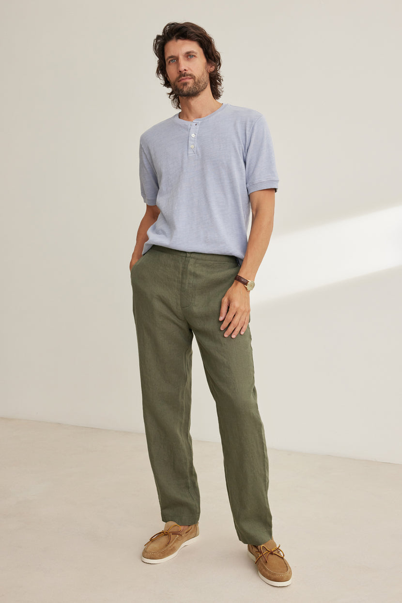A man stands indoors with his hands in his pockets, wearing a light blue short-sleeve henley shirt, tan boat shoes, and Velvet by Graham & Spencer ALFRED LINEN PANT—straight-leg olive green pants with an elastic waistband—against a plain background.