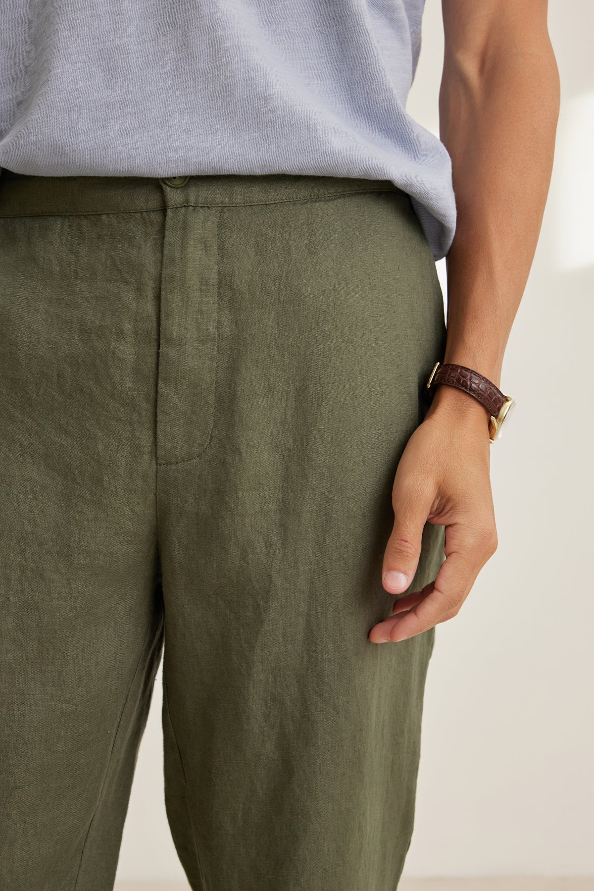 A person wearing Velvet by Graham & Spencer's ALFRED LINEN PANT in olive green with an elastic waistband, paired with a light gray shirt and a brown leather wristwatch, stands with one hand by their side.-39152330670273
