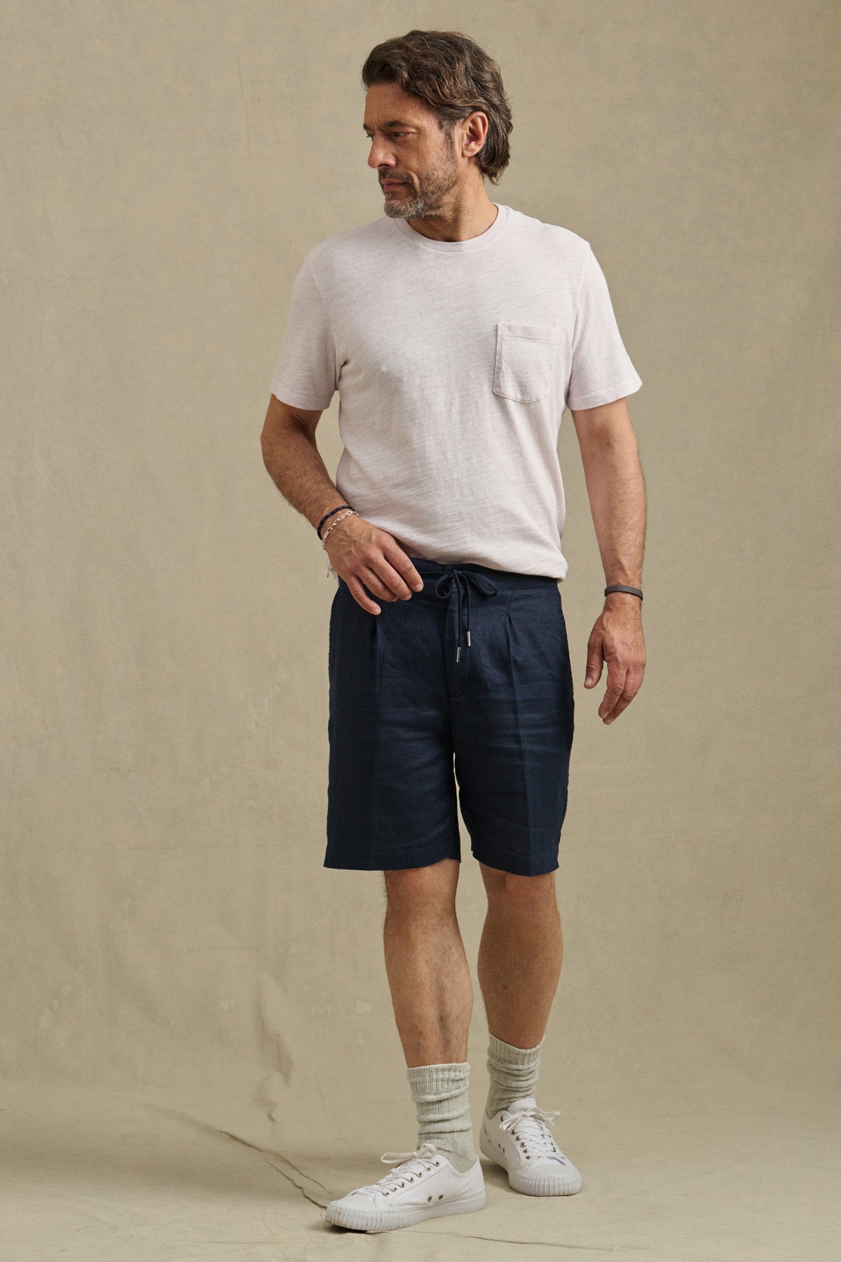 A man stands against a plain beige background, wearing the Velvet by Graham & Spencer DAMIEN LINEN SHORT with a light T-shirt, socks, and white sneakers.