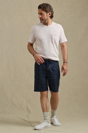 A man stands against a plain beige background, wearing the Velvet by Graham & Spencer DAMIEN LINEN SHORT with a light T-shirt, socks, and white sneakers.