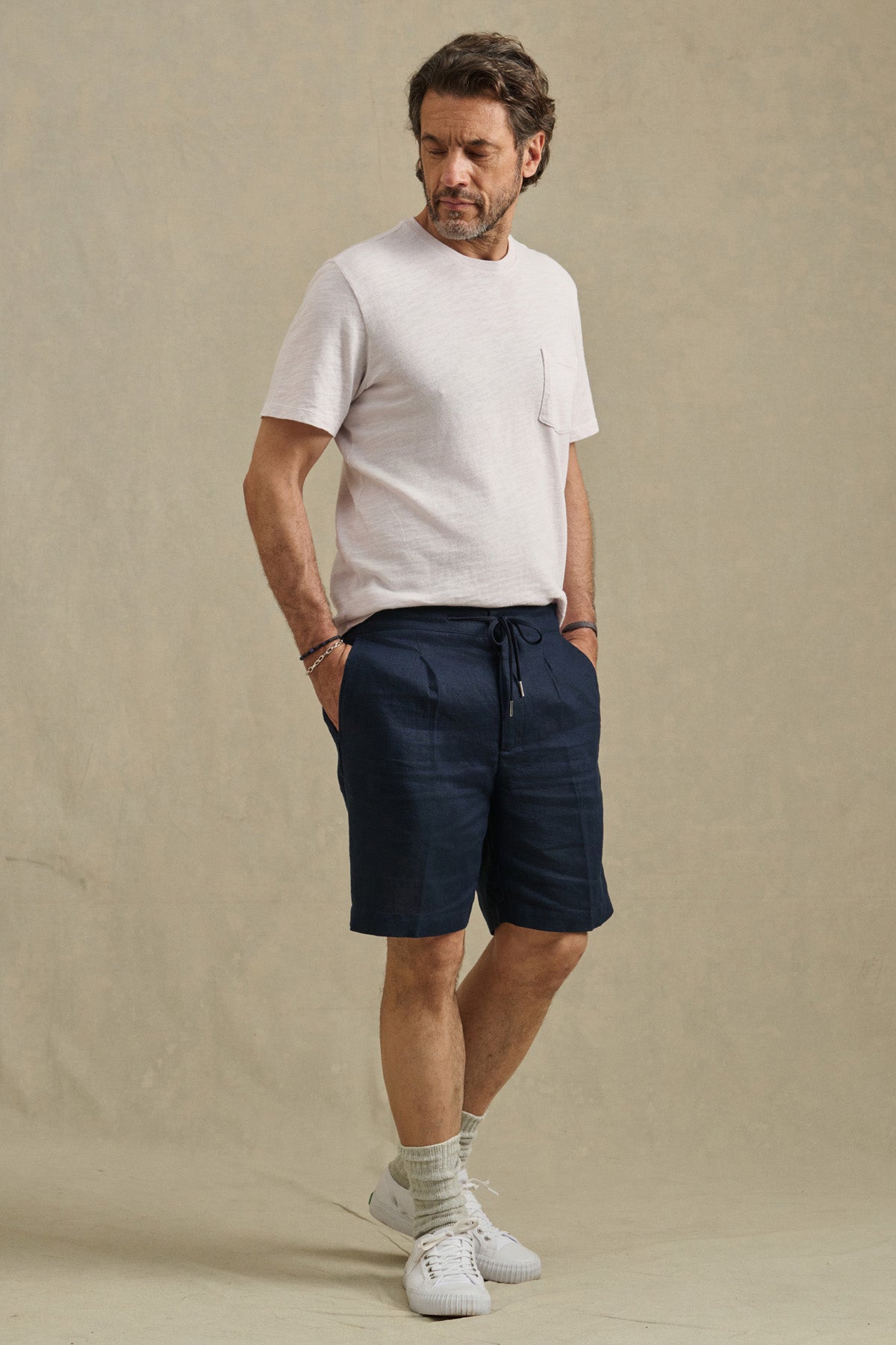 A man stands against a neutral backdrop wearing the DAMIEN LINEN SHORT by Velvet by Graham & Spencer, paired with a light short-sleeve t-shirt, light socks, and white sneakers, his hands in his pockets.