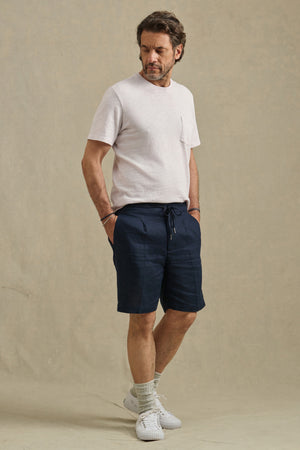 A man stands against a neutral backdrop wearing the DAMIEN LINEN SHORT by Velvet by Graham & Spencer, paired with a light short-sleeve t-shirt, light socks, and white sneakers, his hands in his pockets.