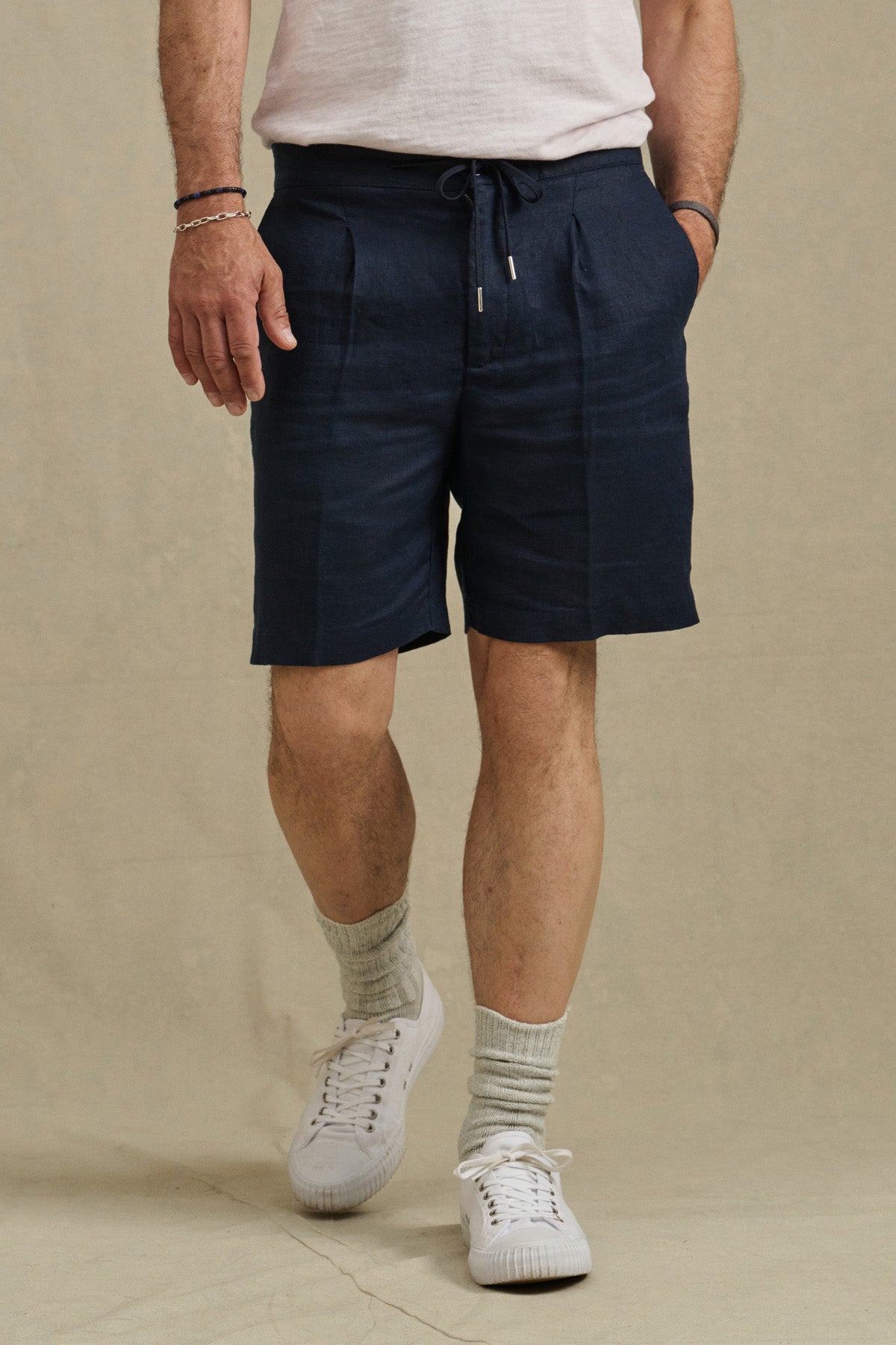 Wearing Velvet by Graham & Spencer’s DAMIEN LINEN SHORT in navy, a white tee, white socks, and sneakers, a person stands against a plain beige background.
