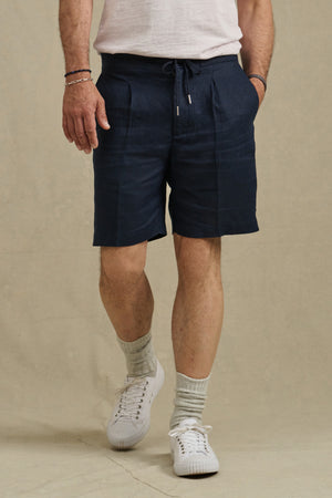 Wearing Velvet by Graham & Spencer’s DAMIEN LINEN SHORT in navy, a white tee, white socks, and sneakers, a person stands against a plain beige background.
