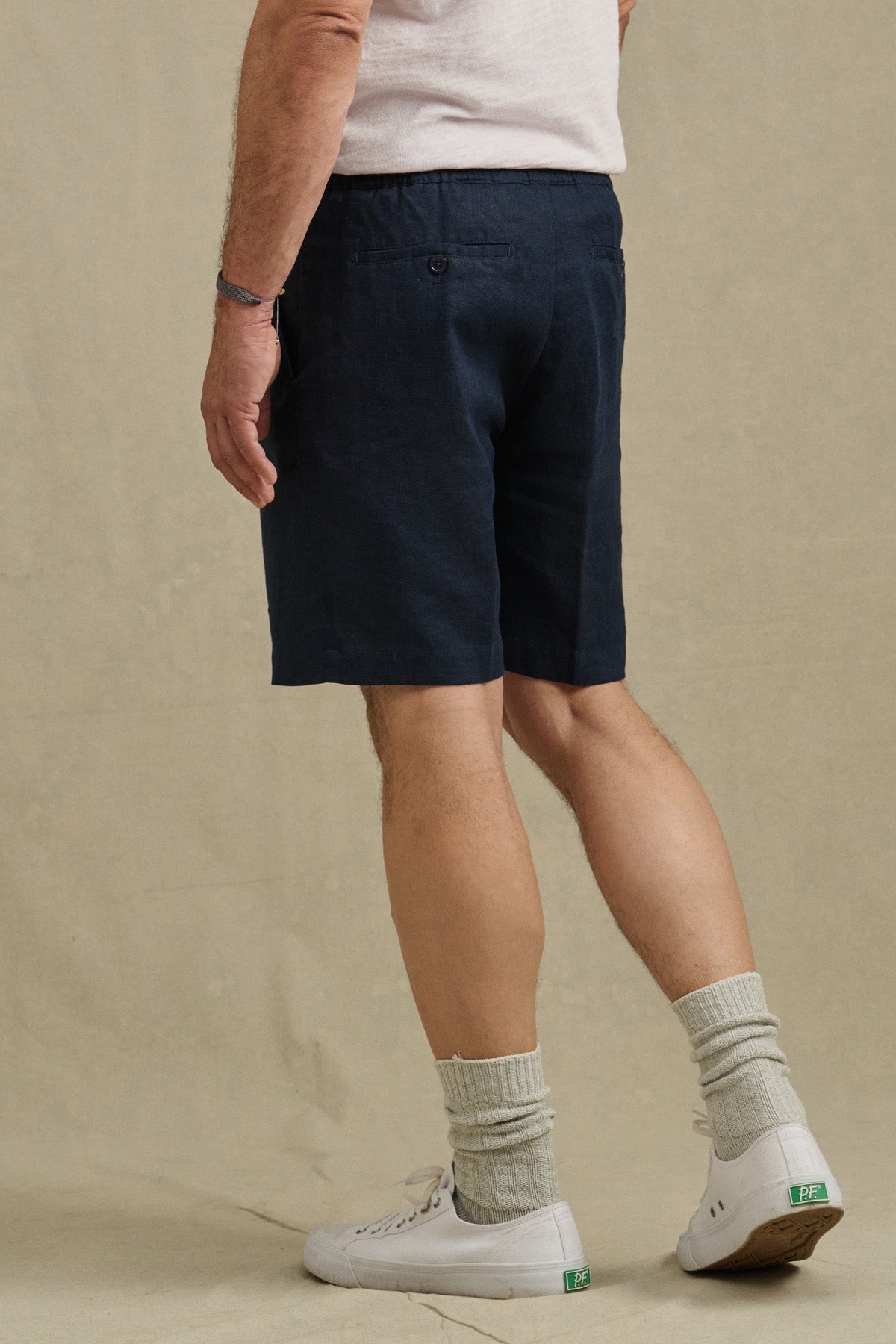 A person is seen from behind wearing a white t-shirt, DAMIEN LINEN SHORT by Velvet by Graham & Spencer in navy blue, light gray socks, and white sneakers.
