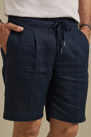 A person wearing Velvet by Graham & Spencer's DAMIEN LINEN SHORT in dark blue, with hands in pockets, is photographed from waist to mid-thigh.