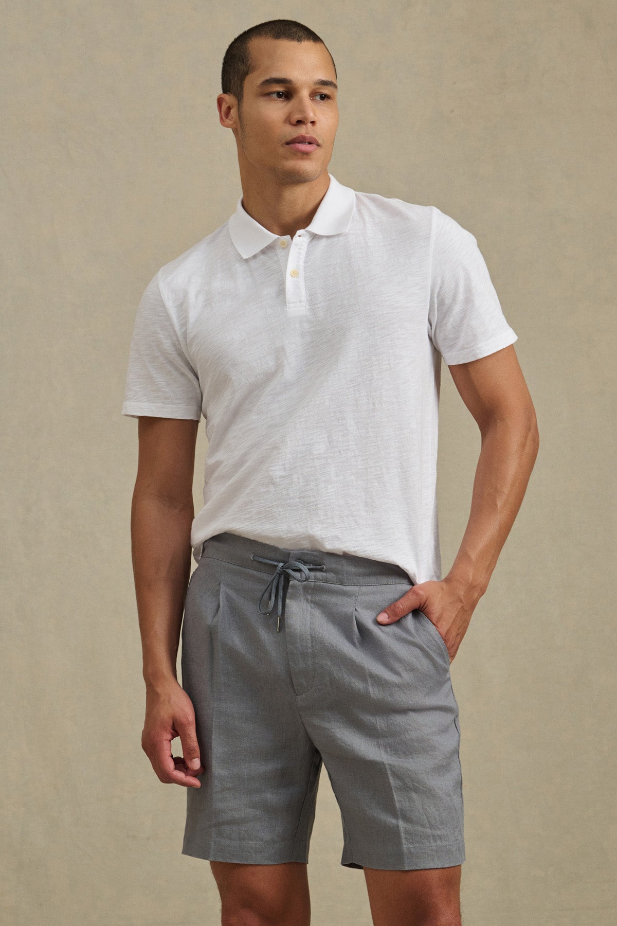 A man stands against a plain beige background wearing light gray DAMIEN LINEN SHORTS by Velvet by Graham & Spencer and a white short-sleeve polo shirt.-41374384423105