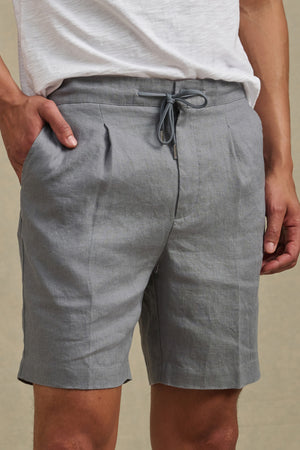 A person wearing Velvet by Graham & Spencer’s DAMIEN LINEN SHORT in light gray with front pleats and drawstring, paired with a white T-shirt, stands with one hand in their pocket.