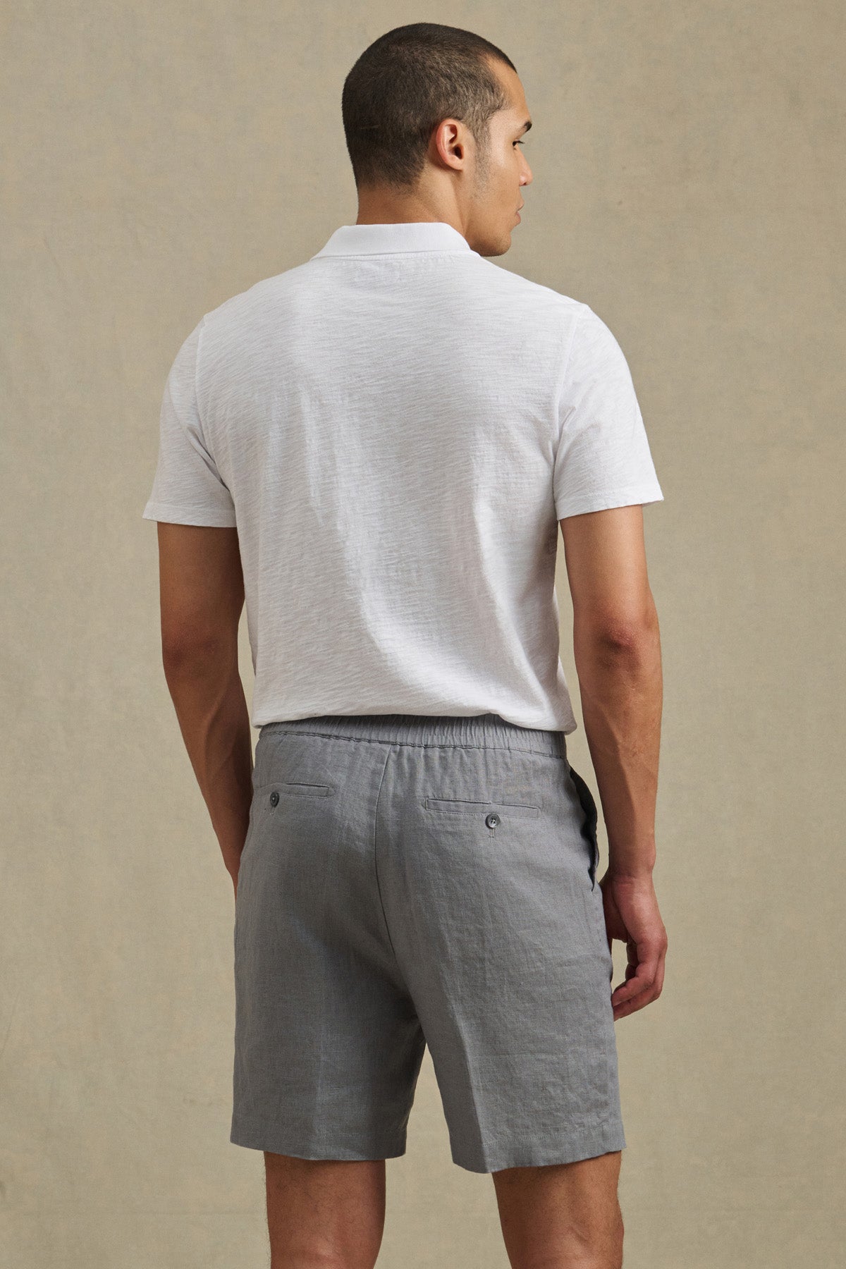 A man, seen from behind against a neutral background, wears the DAMIEN LINEN SHORT by Velvet by Graham & Spencer with a white short-sleeve polo shirt and gray shorts.