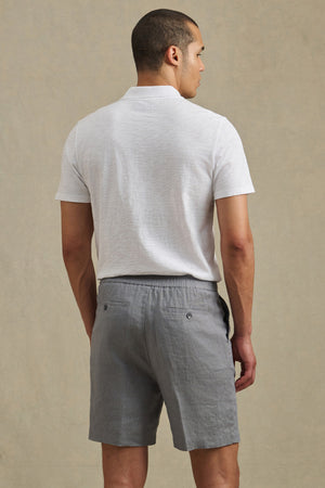 A man, seen from behind against a neutral background, wears the DAMIEN LINEN SHORT by Velvet by Graham & Spencer with a white short-sleeve polo shirt and gray shorts.