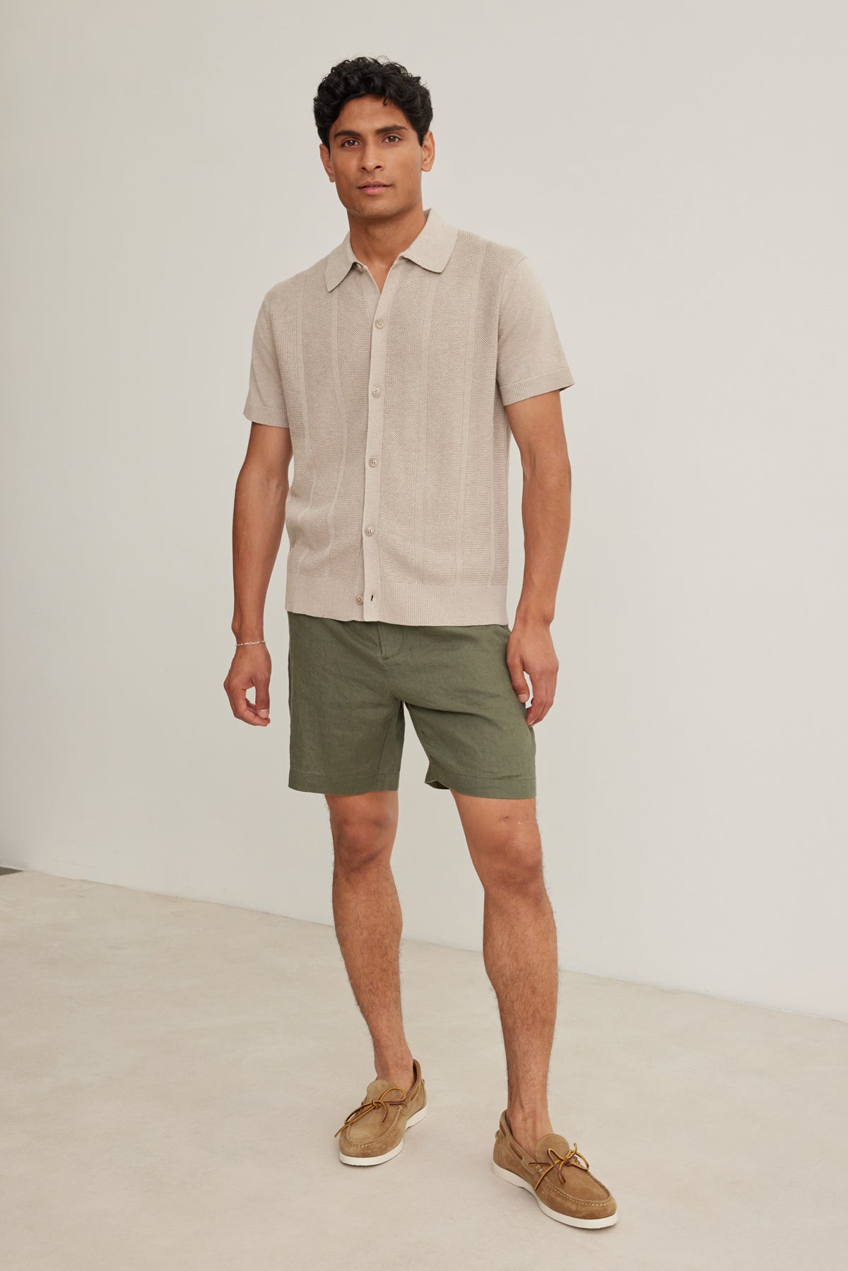 A man stands indoors against a plain light background wearing Velvet by Graham & Spencer's EAMON LINEN SHORT, paired with a beige linen short-sleeve shirt and tan boat shoes—a versatile, stylish look for warm weather.-39152684695745