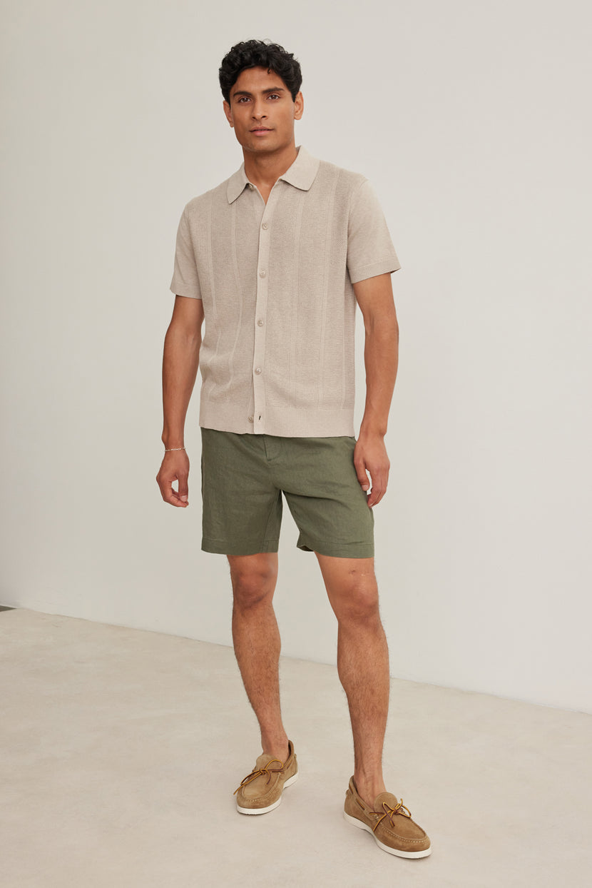 A man stands indoors against a plain light background wearing Velvet by Graham & Spencer's EAMON LINEN SHORT, paired with a beige linen short-sleeve shirt and tan boat shoes—a versatile, stylish look for warm weather.
