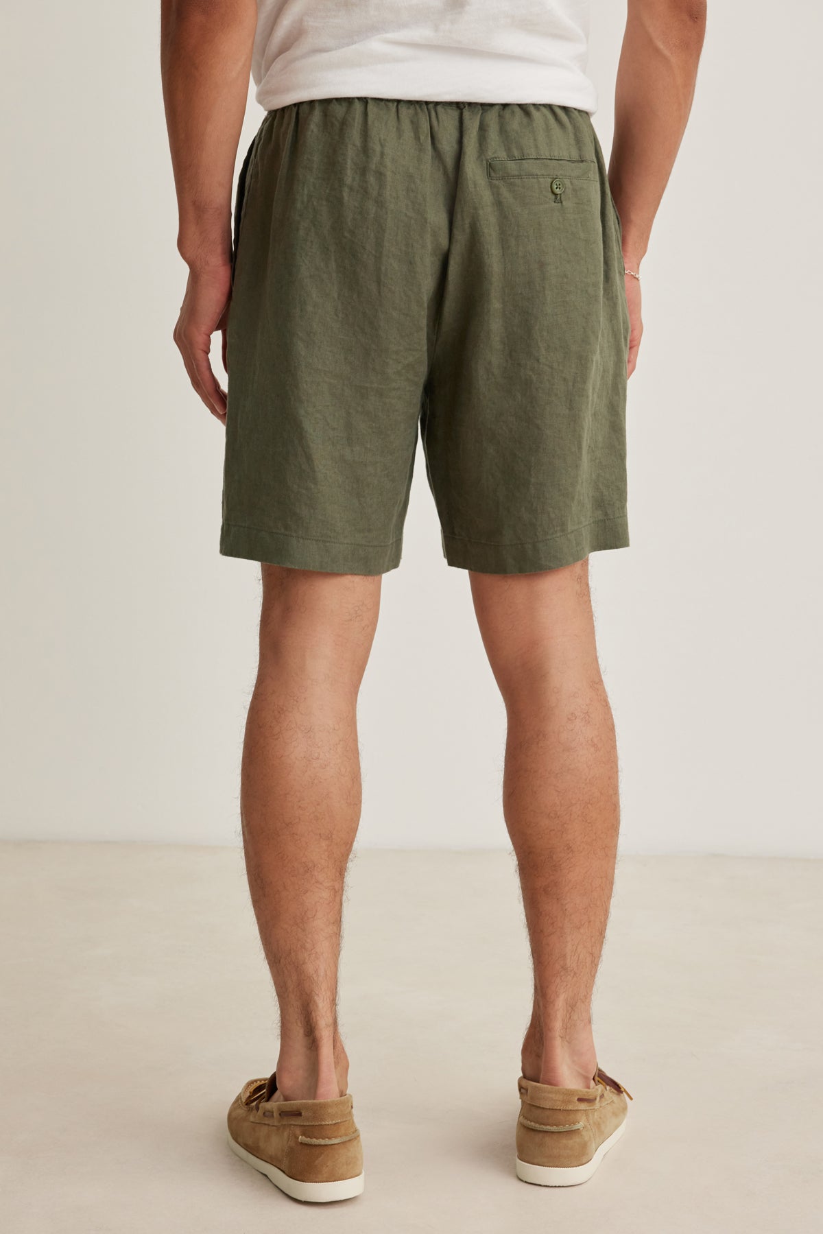 A person stands indoors, shown from the back, wearing a white shirt, brown loafers, and Velvet by Graham & Spencer's EAMON LINEN SHORT—an ideal outfit for warm weather and effortless style.-39152684597441