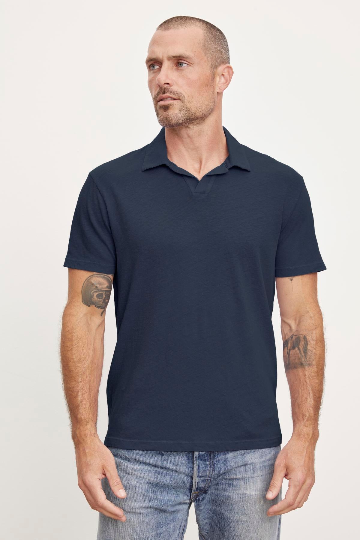   A man in a navy Velvet by Graham & Spencer BECK Polo shirt and jeans stands facing forward, with visible tattoos on both forearms. 