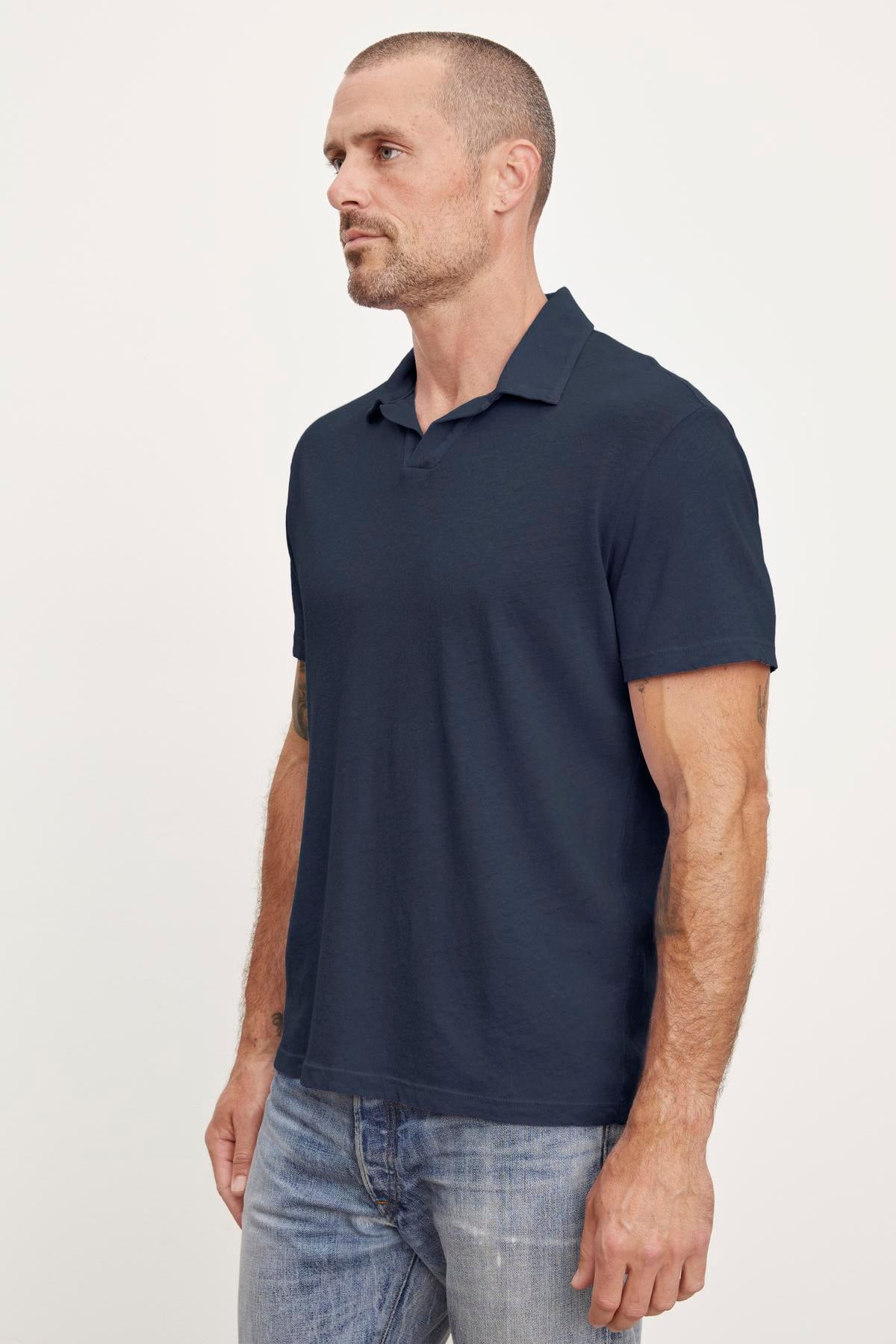   A man in a navy blue Velvet by Graham & Spencer BECK POLO linen blend polo shirt and jeans, standing in a profile view with a neutral expression. 