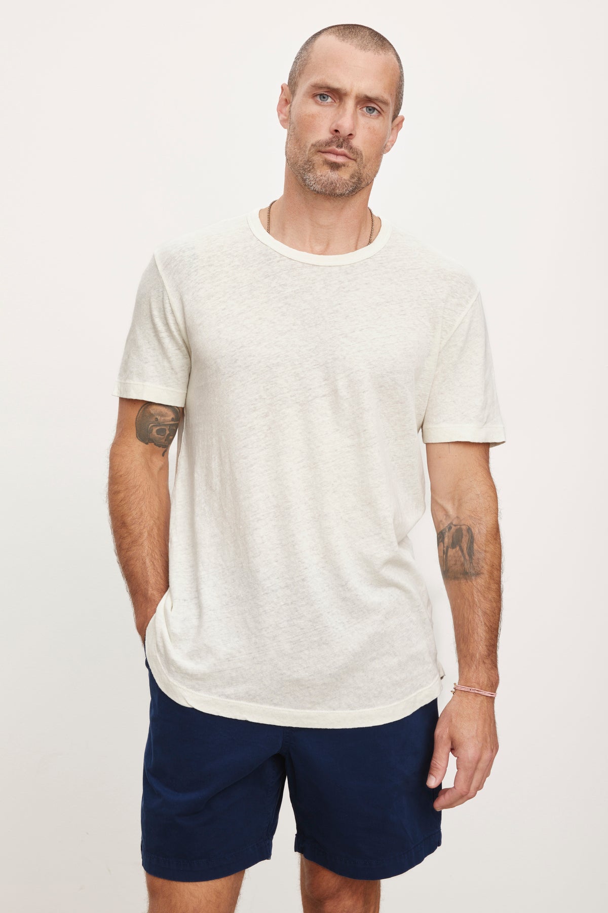  A man with a beard and tattoos stands against a white background, wearing a Velvet by Graham & Spencer Davey Tee in light gray and navy shorts. 