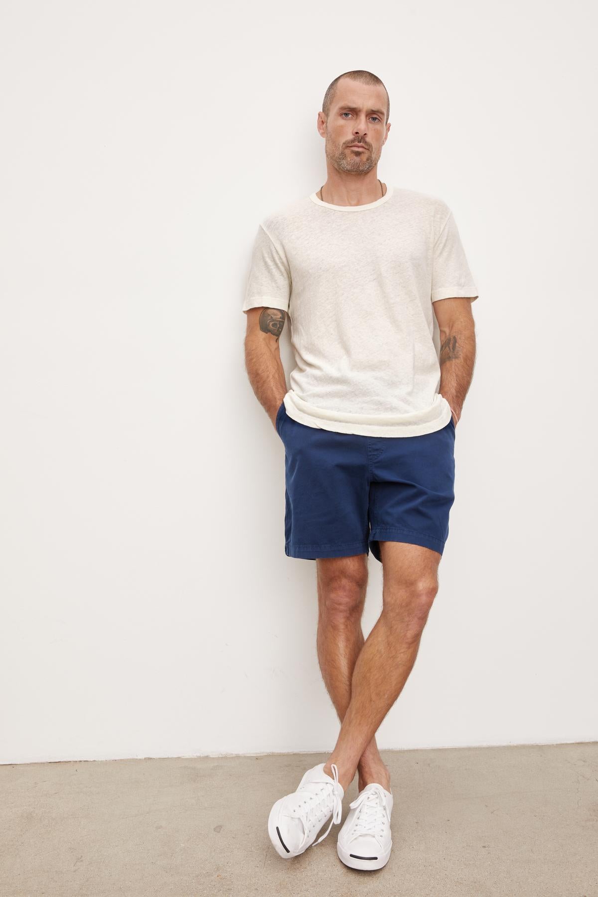   A man standing against a plain background, wearing a Velvet by Graham & Spencer Davey Tee, blue shorts, and white sneakers, with his right leg crossed over his left. 