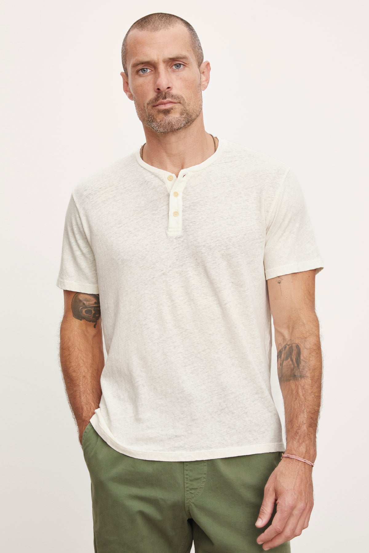   A man with a short beard and tattoos on his arms wearing a Velvet by Graham & Spencer Lionel Henley style shirt and olive green pants, standing against a white background. 