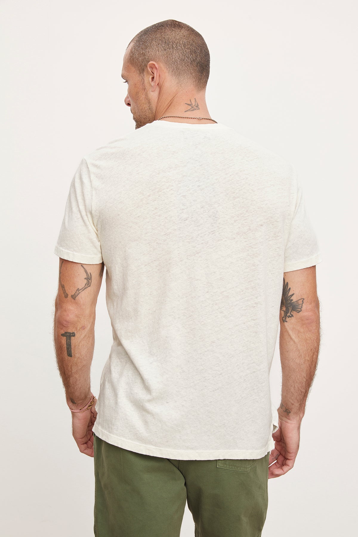   Man standing with back to the camera, showcasing various tattoos on his arms and neck, wearing a white Velvet by Graham & Spencer henley style t-shirt and green trousers. 