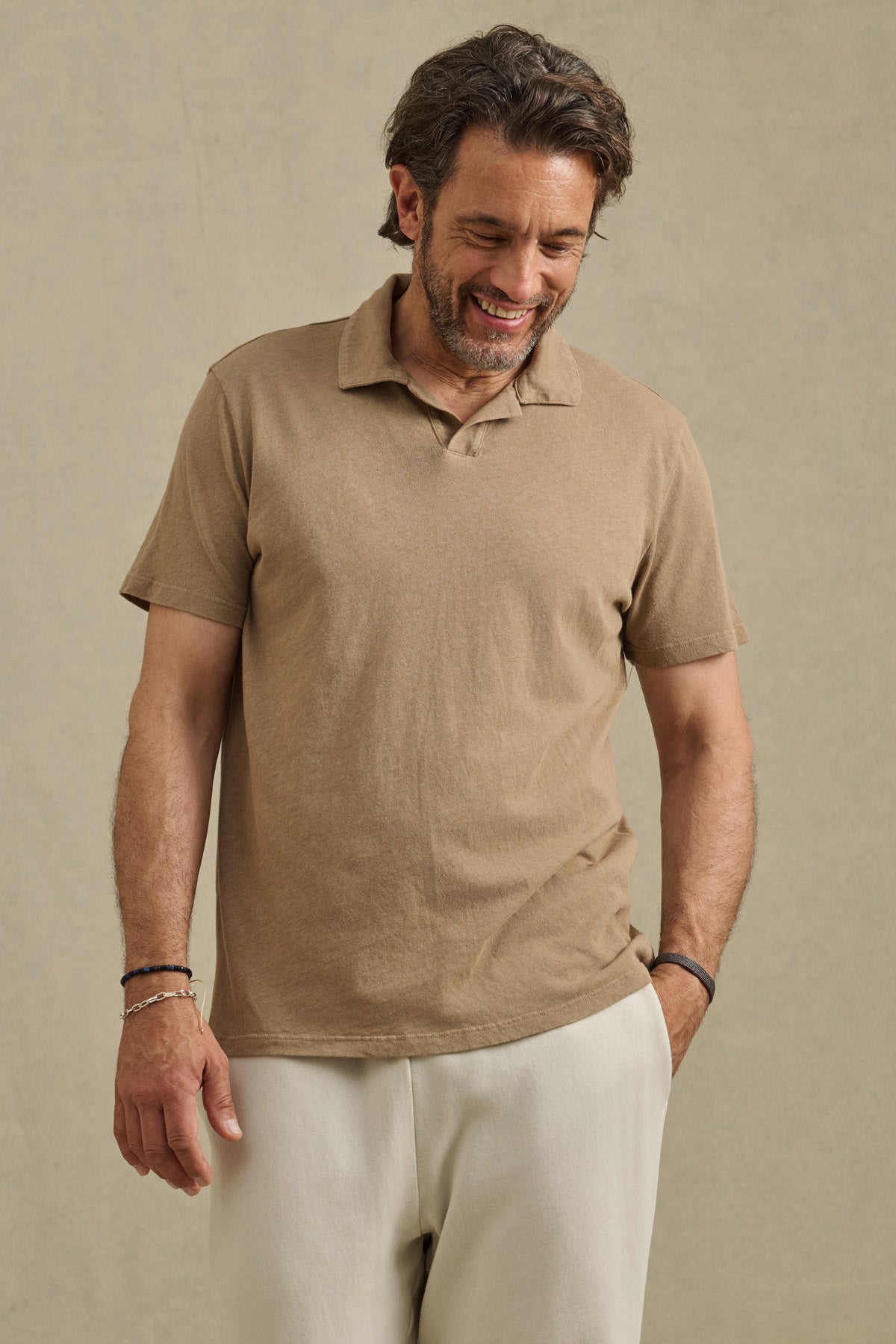 A man with medium-length hair and a beard, smiling with one hand in his pocket, models the relaxed fit ASHTON POLO by Velvet by Graham & Spencer, paired with light pants against a plain background.