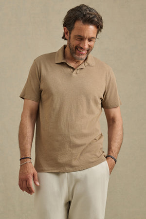A man with medium-length hair and a beard, smiling with one hand in his pocket, models the relaxed fit ASHTON POLO by Velvet by Graham & Spencer, paired with light pants against a plain background.