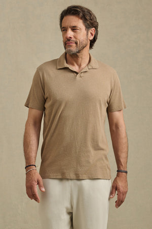 A man with short hair and a beard wears the ASHTON POLO by Velvet by Graham & Spencer— a tan, relaxed fit cotton-linen blend—paired with light pants, standing against a plain beige background.