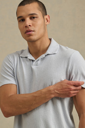 A man wearing the lightweight ASHTON POLO by Velvet by Graham & Spencer stands against a neutral background, touching his upper arm with one hand.