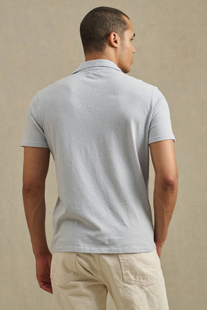 A man, seen from behind, wears the ASHTON POLO by Velvet by Graham & Spencer—a light gray cotton-linen short-sleeve shirt—paired with off-white pants, set against a neutral background.