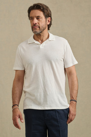 A bearded man wears the Velvet by Graham & Spencer ASHTON POLO in white, styled with dark pants, standing against a plain light background.