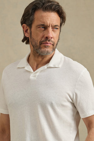 A man with medium-length brown hair and a beard models the relaxed-fit ASHTON POLO by Velvet by Graham & Spencer, gazing slightly to the side against a neutral background.
