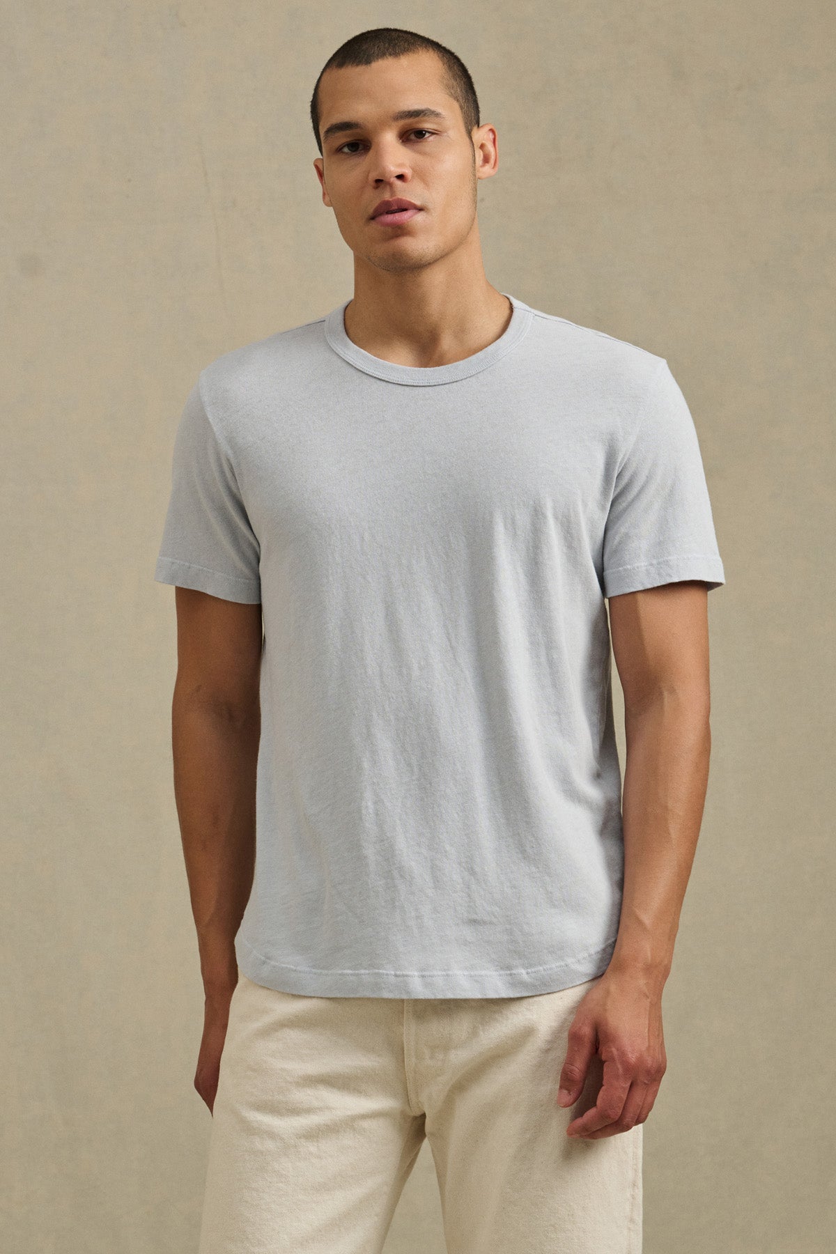 A person stands against a neutral background wearing the Velvet by Graham & Spencer LUKA TEE, a light gray cotton-linen crew neck short-sleeve t-shirt, paired with off-white pants and looking directly at the camera.-41479919206593