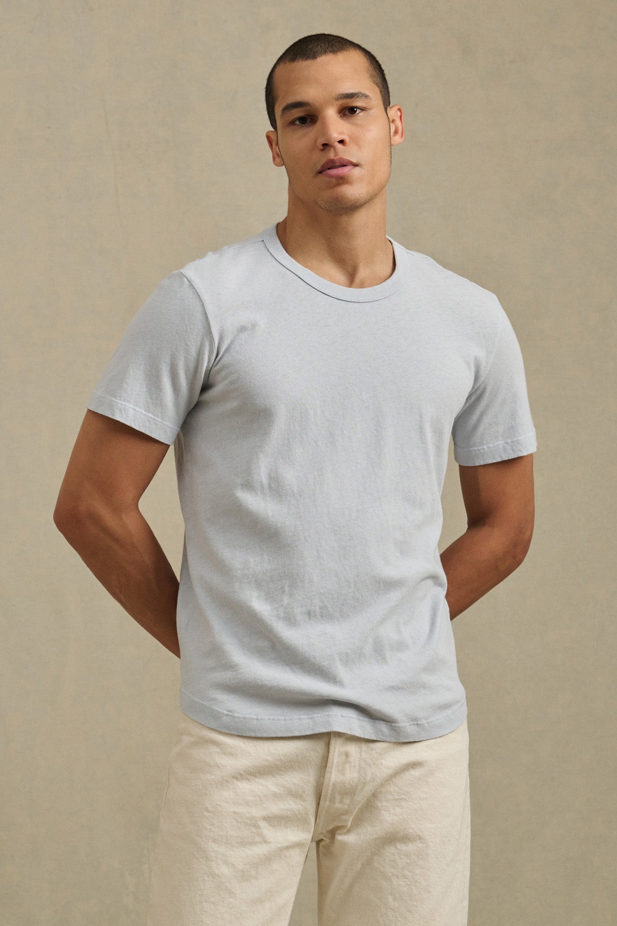 A person stands against a plain beige background, wearing the LUKA TEE by Velvet by Graham & Spencer with off-white pants, hands behind their back.-41479919272129