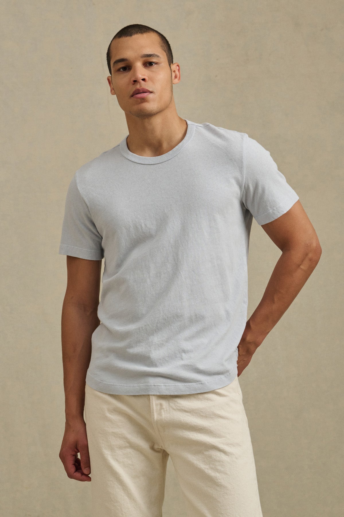 A person wears the Velvet by Graham & Spencer LUKA TEE and off-white pants, standing against a plain beige background.