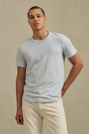 A person wears the Velvet by Graham & Spencer LUKA TEE and off-white pants, standing against a plain beige background.