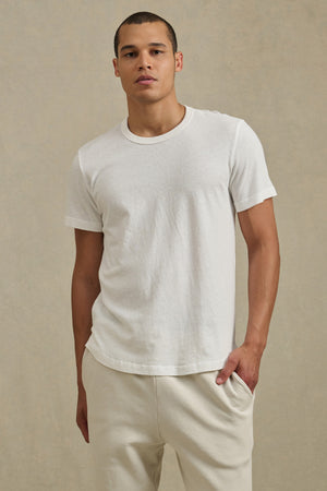 Wearing the LUKA TEE by Velvet by Graham & Spencer, styled with cream-colored cotton-linen pants, a person stands against a neutral beige background with one hand in their pocket.