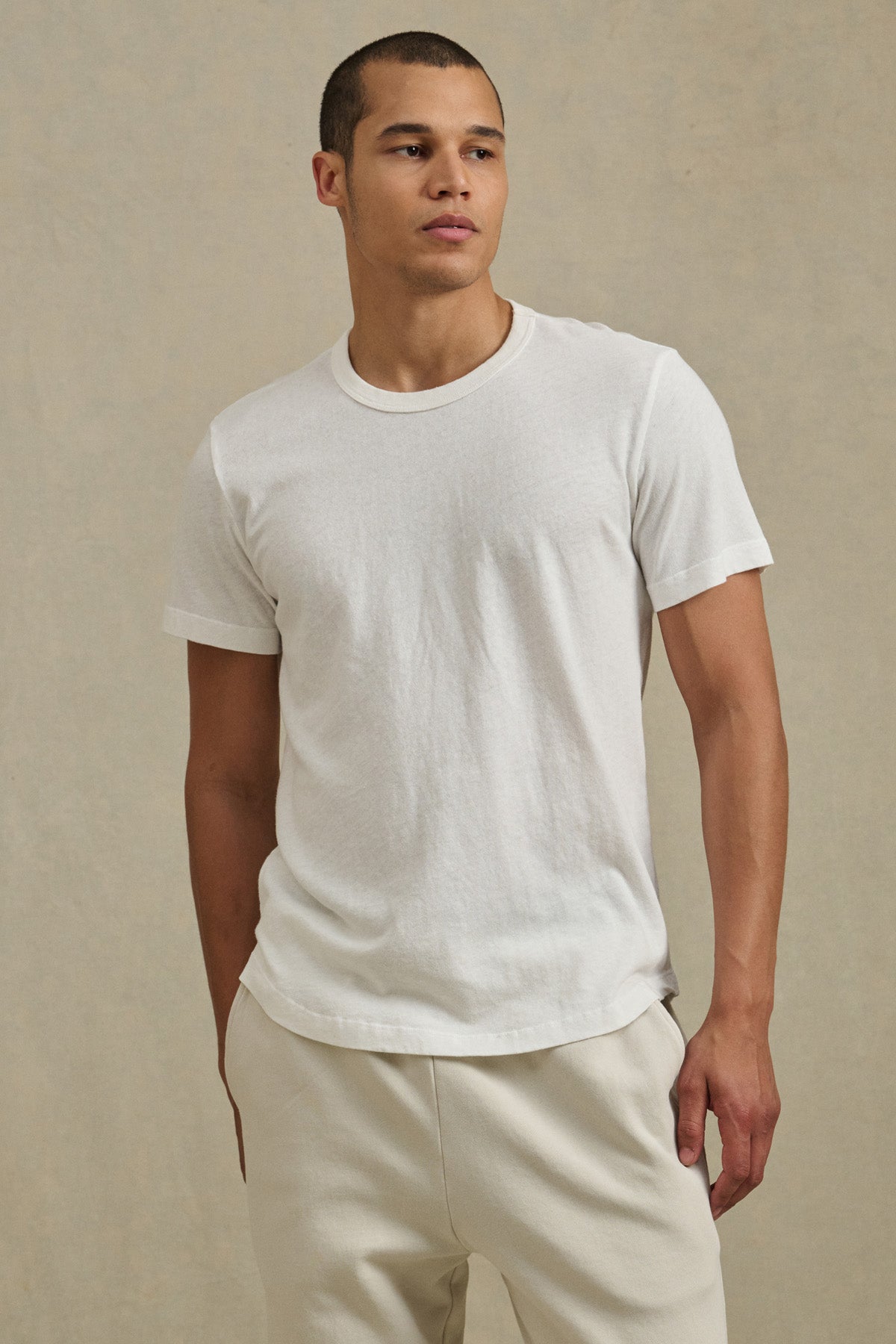 A man wears the LUKA TEE by Velvet by Graham & Spencer—a plain white crew neck short-sleeve cotton-linen shirt—paired with light pants, standing against a beige background for an effortless warm-weather look.