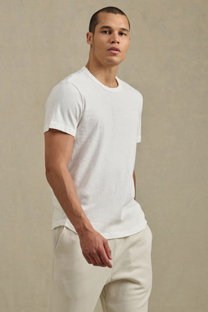 A person stands against a neutral background wearing the LUKA TEE by Velvet by Graham & Spencer—a lightweight cotton-linen crew neck white t-shirt—paired with light pants, looking slightly to the side for a casual, polished look.