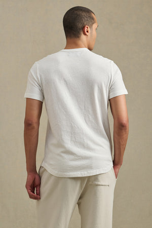 A person stands facing away from the camera, wearing the LUKA TEE by Velvet by Graham & Spencer, a white crew neck short-sleeve shirt crafted from lightweight cotton-linen—ideal for warm weather—paired with light pants against a neutral background.