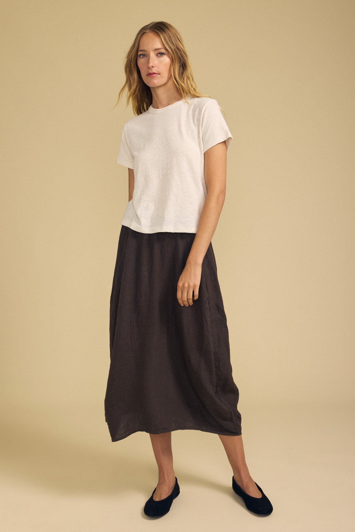 A person stands against a beige background wearing a white short-sleeve t-shirt, black flats, and the FAE LINEN SKIRT by Velvet by Graham & Spencer—a dark brown, A-line midi skirt.