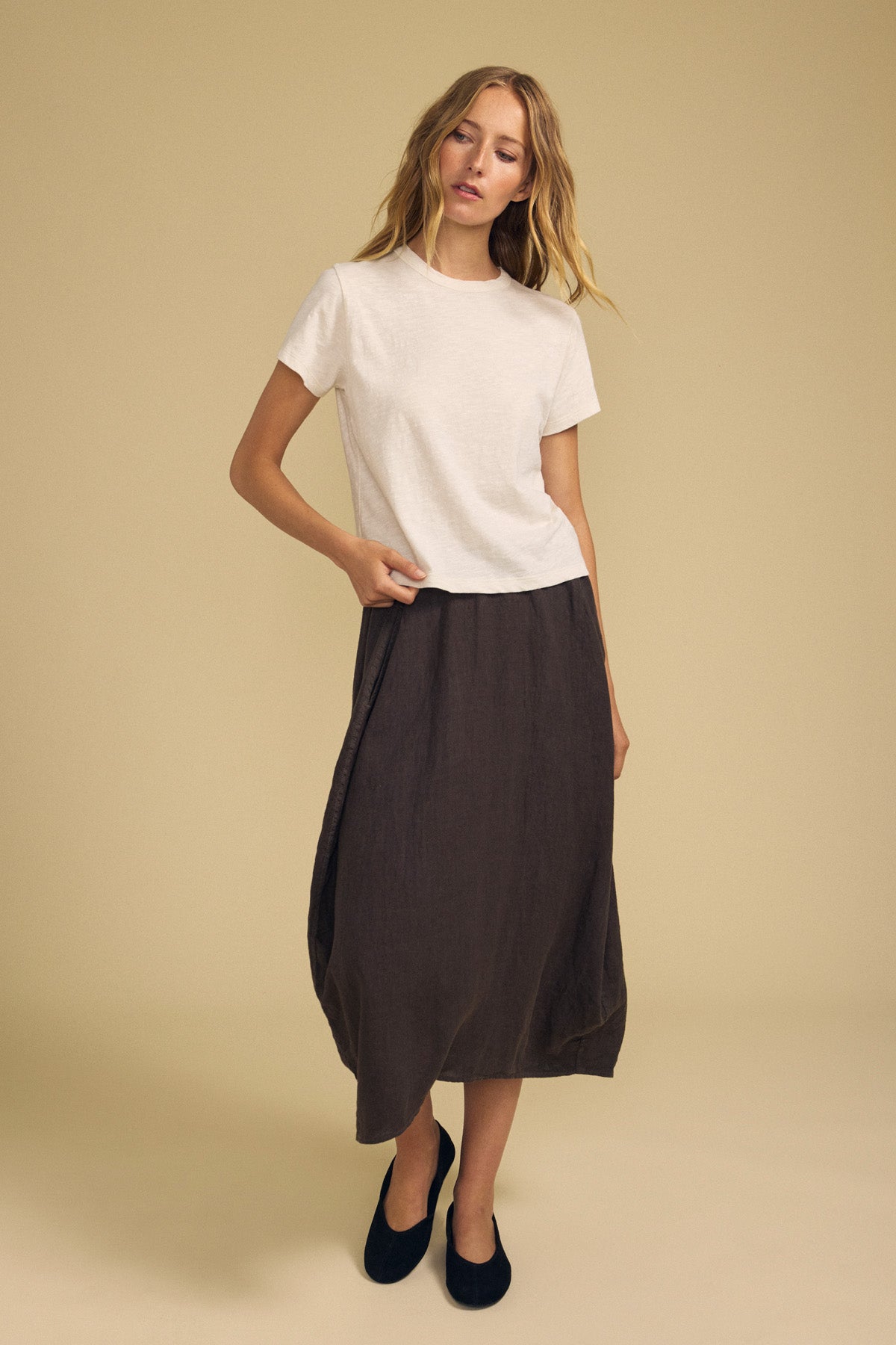 A woman stands against a beige background, wearing a white short-sleeve shirt paired with the FAE LINEN SKIRT by Velvet by Graham & Spencer—a loose, dark brown A-line skirt featuring an elastic waistband—and black shoes.-41960838234305