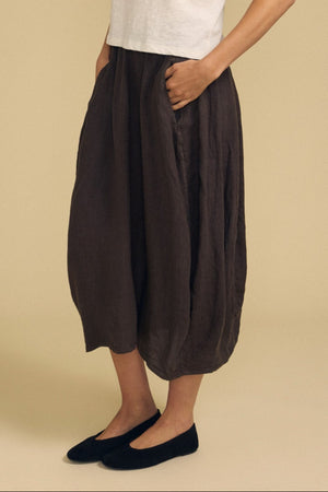 A woman stands against a beige background, wearing the Velvet by Graham & Spencer FAE LINEN SKIRT paired with a white short-sleeve shirt and black shoes.