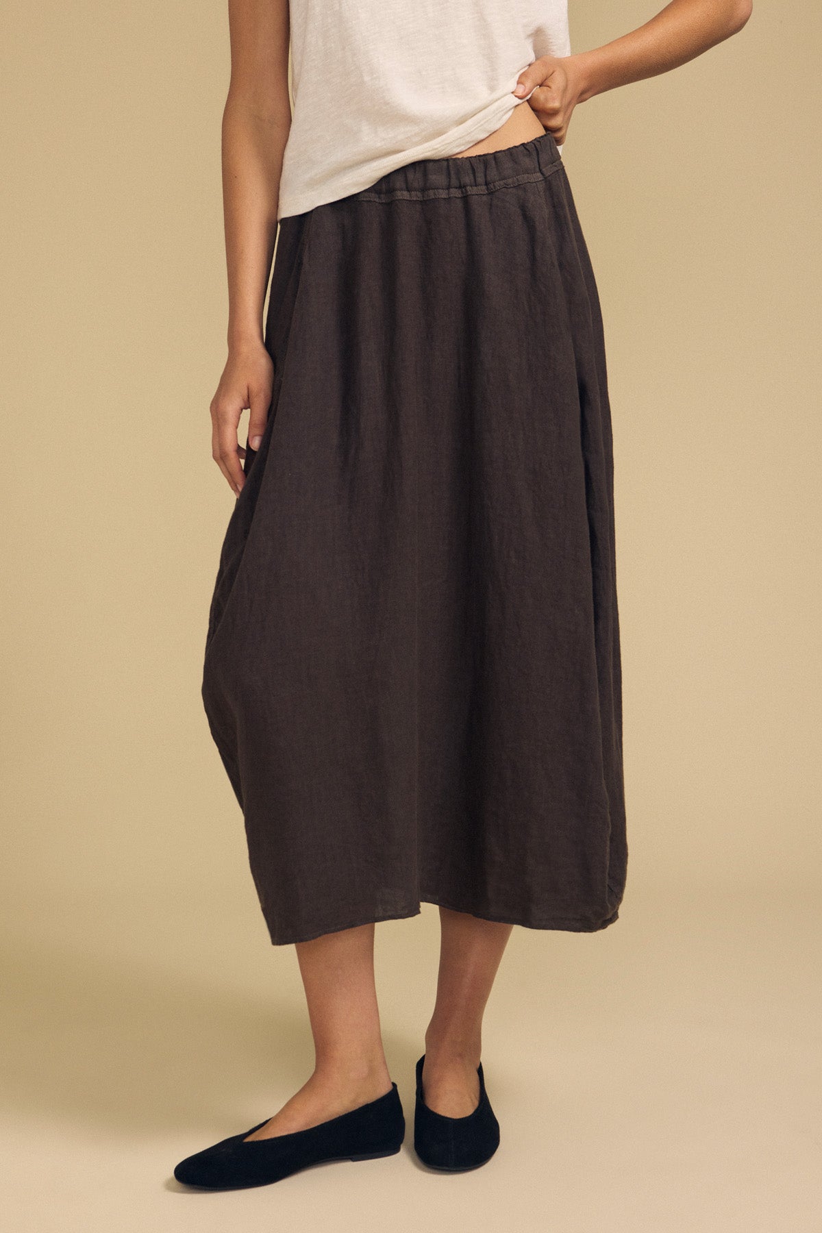 A person wearing a white sleeveless top, the Velvet by Graham & Spencer FAE LINEN SKIRT—a dark brown A-line ankle-length skirt featuring an elastic waistband—and black flats, posing against a plain beige background.-41960838332609