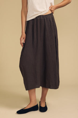 A person wearing a white sleeveless top, the Velvet by Graham & Spencer FAE LINEN SKIRT—a dark brown A-line ankle-length skirt featuring an elastic waistband—and black flats, posing against a plain beige background.