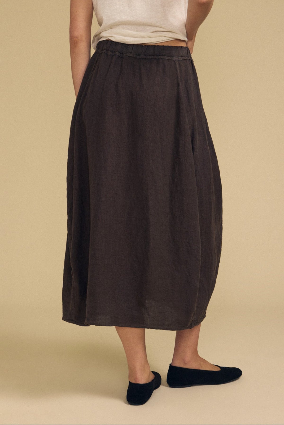 A person stands facing away, wearing a white short-sleeve shirt paired with the FAE LINEN SKIRT by Velvet by Graham & Spencer—a loose dark brown skirt with an elastic waistband and subtle A-line silhouette—finished with black flats against a beige background.-41960838398145