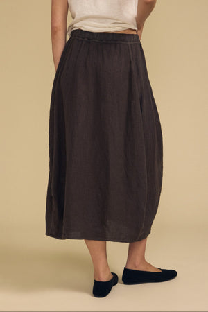 A person stands facing away, wearing a white short-sleeve shirt paired with the FAE LINEN SKIRT by Velvet by Graham & Spencer—a loose dark brown skirt with an elastic waistband and subtle A-line silhouette—finished with black flats against a beige background.