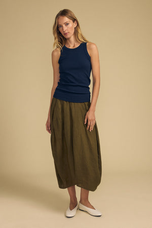 A woman stands against a beige background wearing a navy sleeveless top, the FAE LINEN SKIRT by Velvet by Graham & Spencer in olive green with an elastic waistband, and white flat shoes.