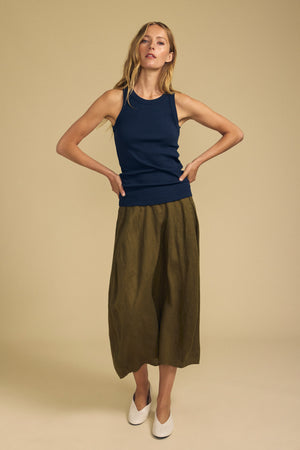 A woman stands against a beige background wearing a navy sleeveless top, the Velvet by Graham & Spencer FAE LINEN SKIRT—an olive green A-line midi skirt with an elastic waistband—and white shoes, her arms relaxed and hands on hips.